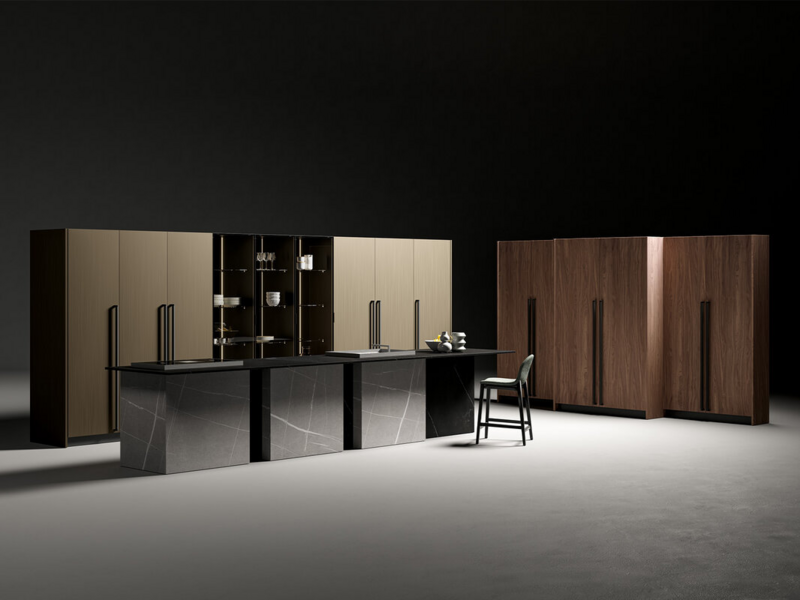 Isola CX 10 laccato opaco nero con maniglia. Top, spalle, schienali e distanziali ceramica Gris Natural Senda e Negro Natural Storm. Colonne CX 10 essenza Canaletto e laccato metallo satinato ottone con maniglia. Colonne vidro CX 19 con struttura laccato opaco nero e vetro grigio fum&egrave;.